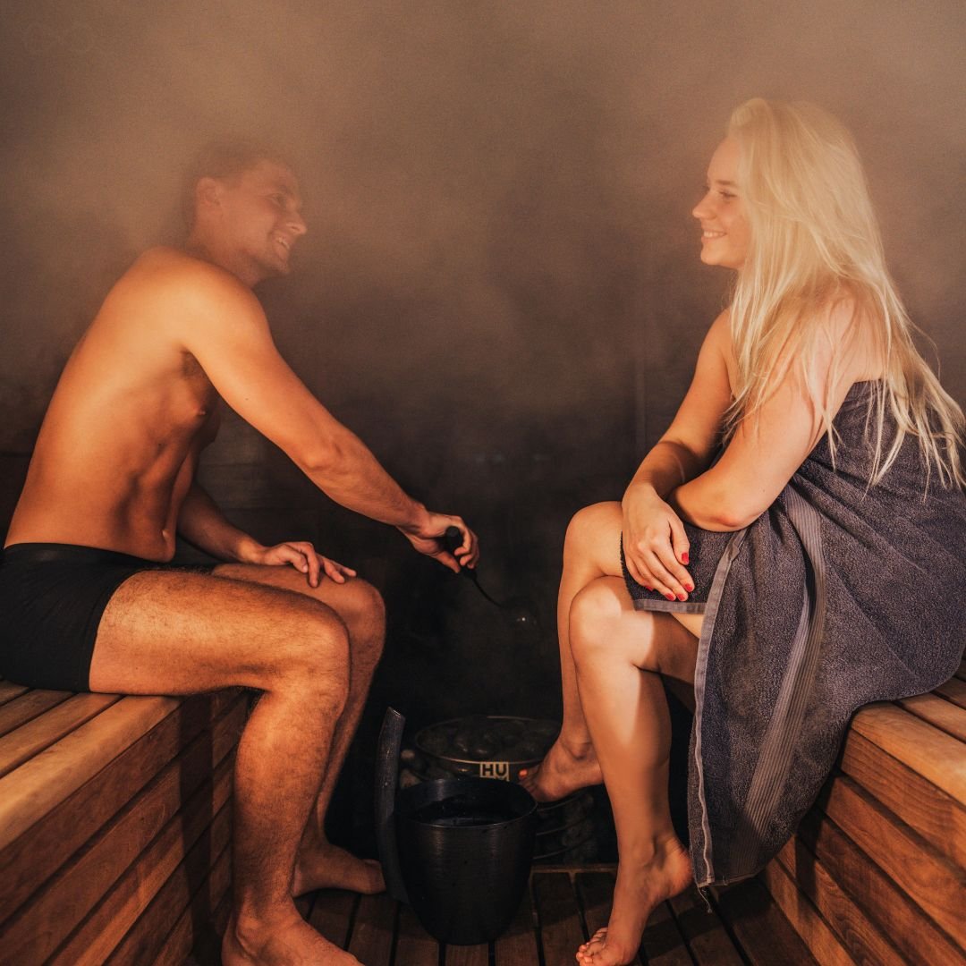 A man and a woman smiling at each other while relaxing in a steamy, wood-paneled sauna. The man is pouring water onto sauna stones with a ladle while the woman sits wrapped in a grey towel.